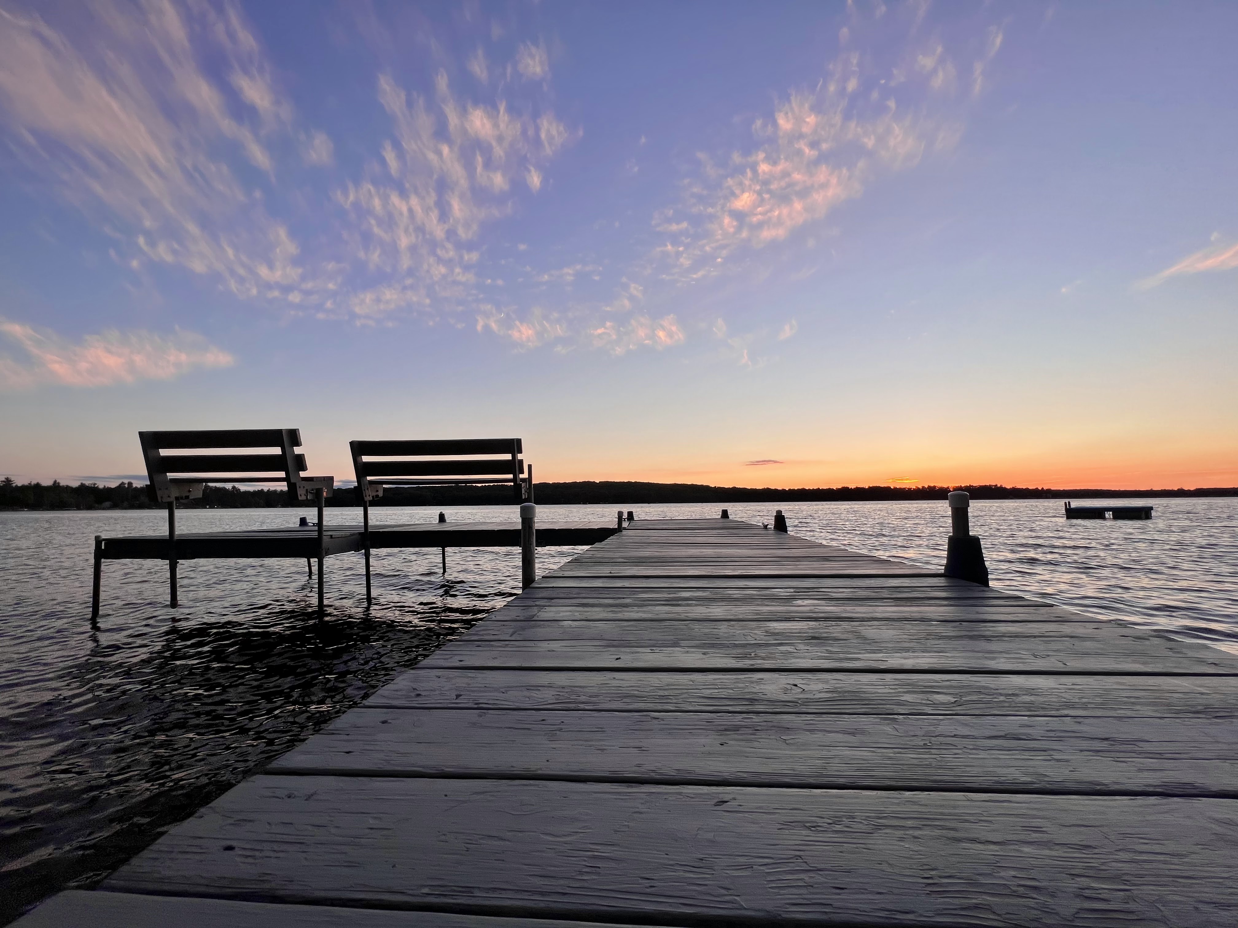 Artisitically shot from the base of an L shaped wooden pier, looking out over the water the pier reaches in front of you with two benches flanking L to the left, their silhouettes standing prominently. The lake water below is choppy. Above the deep blue sky fades to a vibrant orange on the horizon as the sun has already set. A few clouds dot the sky in shades of soft pink.