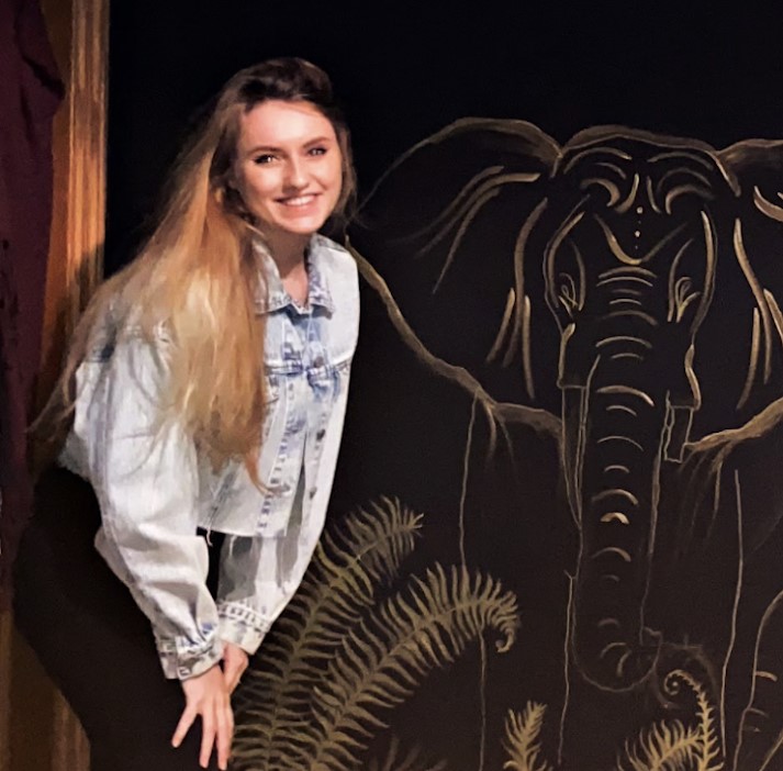 A photo of me, a blonde white woman wearing a lightwashed denim jacket posing in front of a black wall that i hand painted a golden abstract elephant mural on.